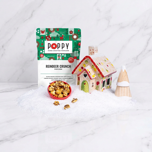 Poppy Handcrafted Popcorn Reindeer Crunch snack bag displayed with a small bowl of wrapped popcorn, a miniature gingerbread house, and a small snowman decoration on a bed of white faux snow against a light blue background.