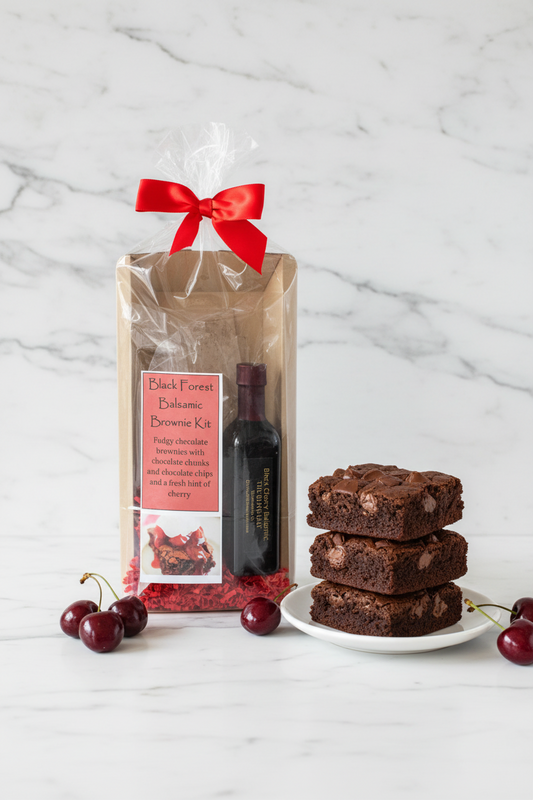 Black Forest Balsamic Brownie Kit in a clear plastic gift bag tied with a red ribbon, containing a bottle of balsamic and chocolate pieces on red paper shred, set against a gray wall and wooden surface.