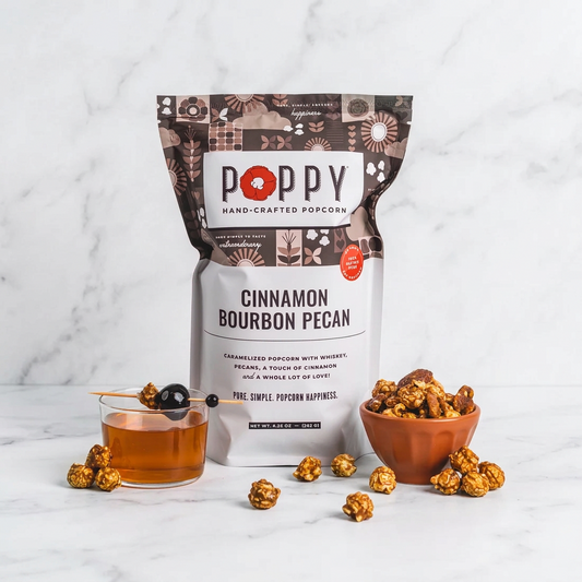 Poppy Cinnamon Bourbon Pecan popcorn package with popcorn and a glass of syrup on a pink background