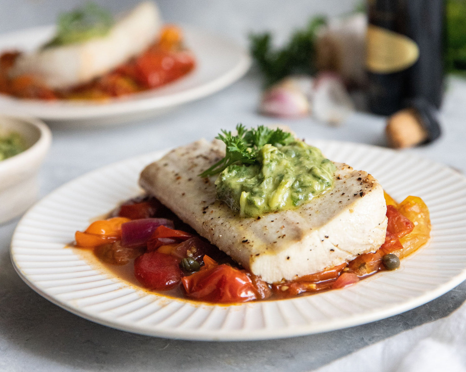 Plated dish of fish with vegetables on a marble surface, with a bottle of wine in the background.