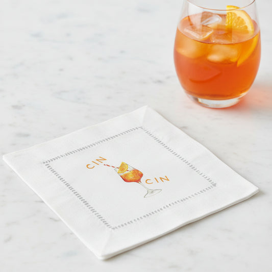 A square white linen napkin featuring a central illustration of an orange cocktail in a glass, with the word 'CIN' repeated above and below, and a gray dotted border around the design.