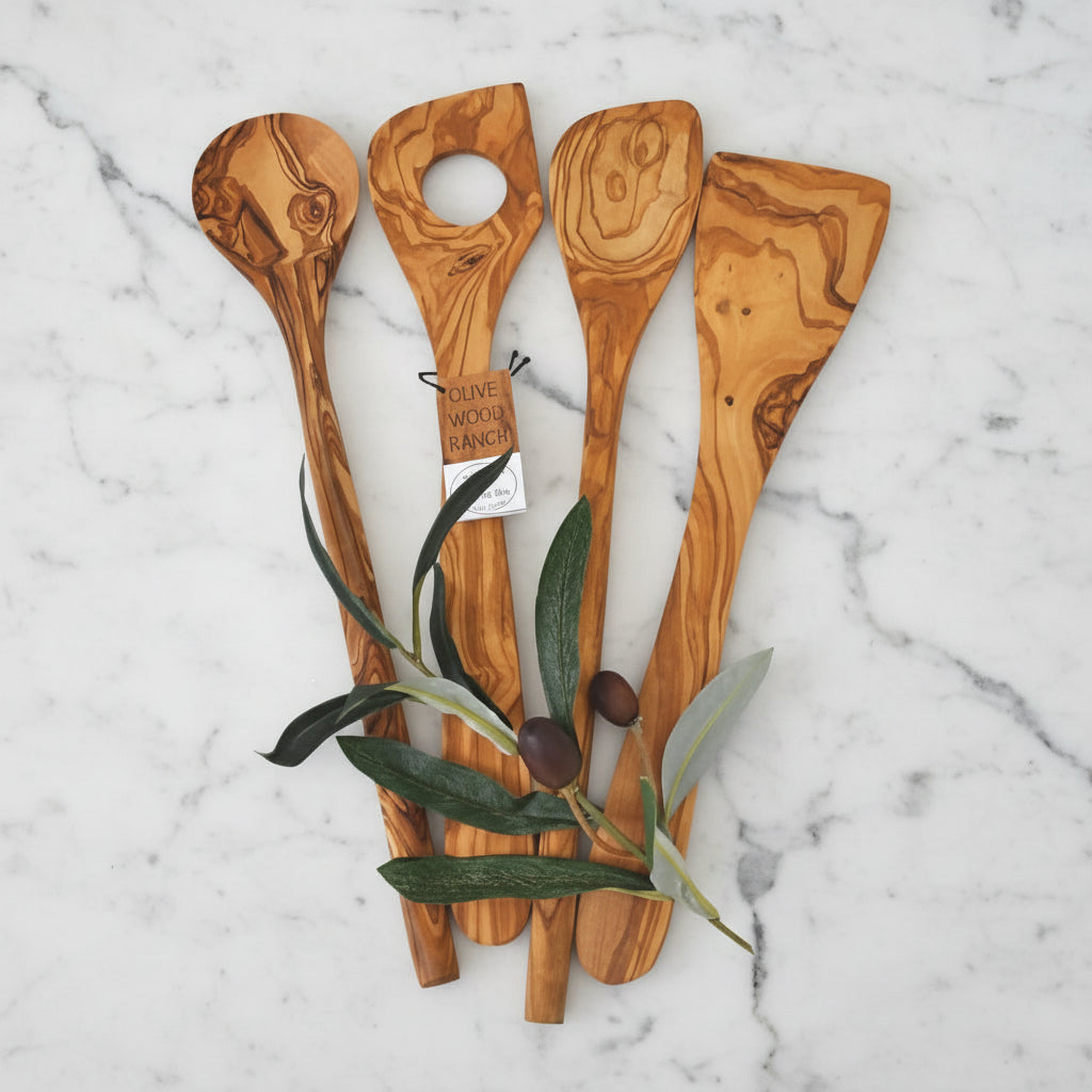 A set of four hand-crafted olive wood kitchen utensils including a spoon, corner spoon, risotto spoon, and spatula, displayed alongside olive leaves.