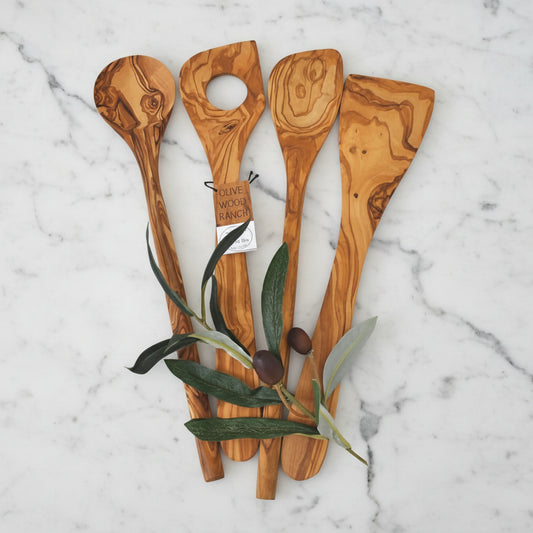 A set of four hand-crafted olive wood kitchen utensils including a spoon, corner spoon, risotto spoon, and spatula, displayed alongside olive leaves.