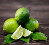 Three whole Tahitian limes with a few leaves on a wooden surface, alongside a sliced lime and a small bowl.