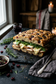 A rustic stuffed focaccia sandwich with cranberries, greens, and melted cheese, served on a dark slate board with a fork, rosemary, and a small bowl of cranberries in a dimly lit kitchen setting.
