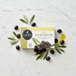 A rectangular box of Lark Fine Foods Olive Scourtin on a light gray background. The packaging is white with a yellow top band, a black circular logo, diagonal yellow stripes on the left, and an image of dark olives on the front.